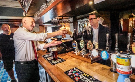Prime minister Andy Burnham hands a pint of beer to chancellor John Healy from behind the bar.